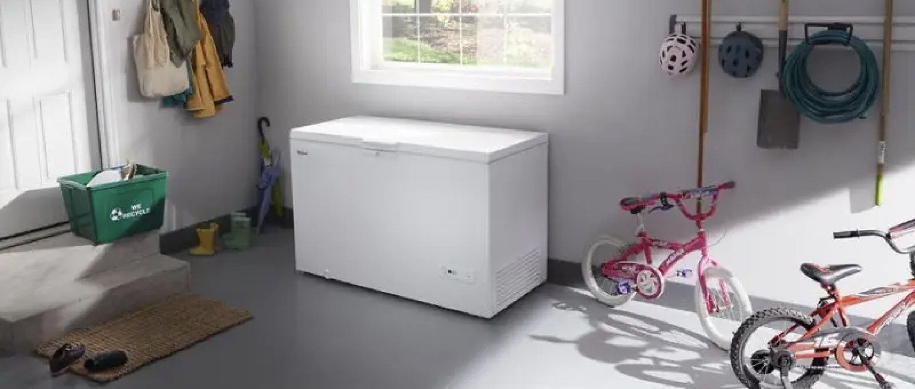 a garage or mudroom with children's bikes and recycling tub, featuring a white chest freezer under a window